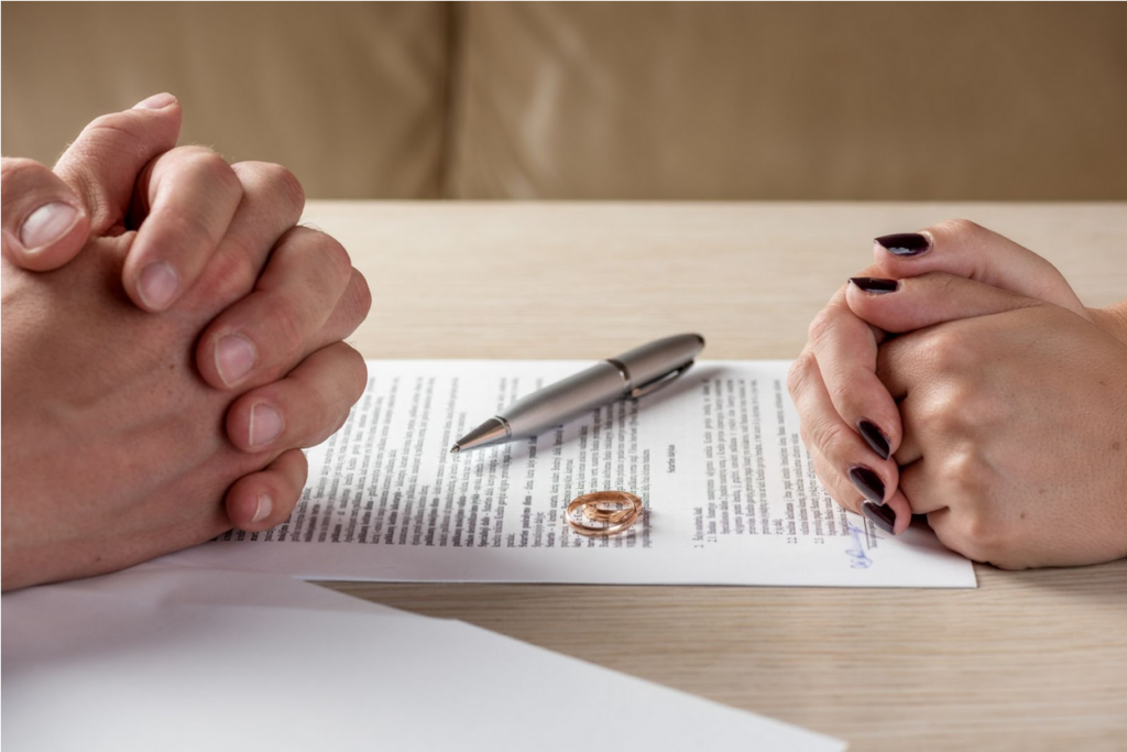 A man and a woman’s hands folded on top of a divorce settlement located on a table. Along with their hands are their wedding rings and a pen on top of the signed paper. 