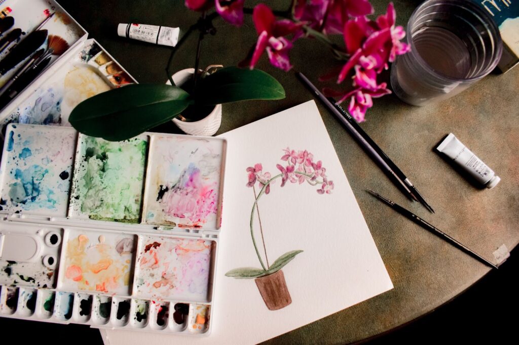 Paints and brushes on a table near a painting of flowers.
