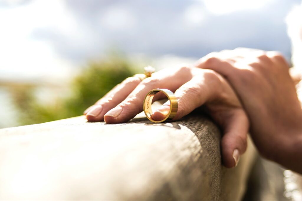 A close-up photo of a woman’s two hands holding onto a railing outside. In her fingers is a man’s wedding ring, off and at her fingertips.
