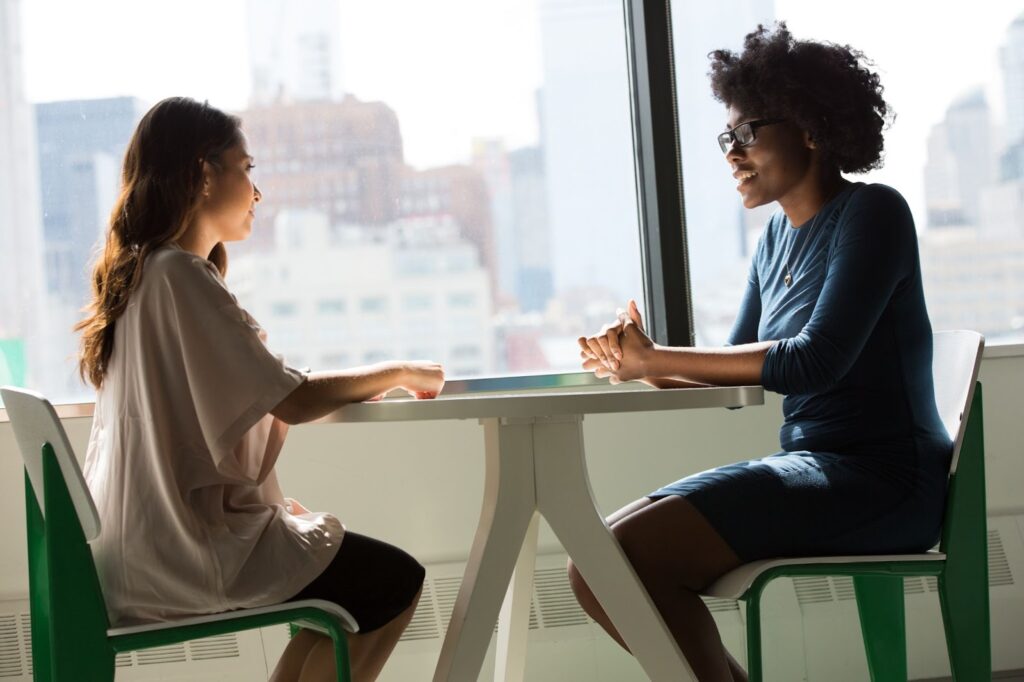 Two women talking together at a table in front of a window.